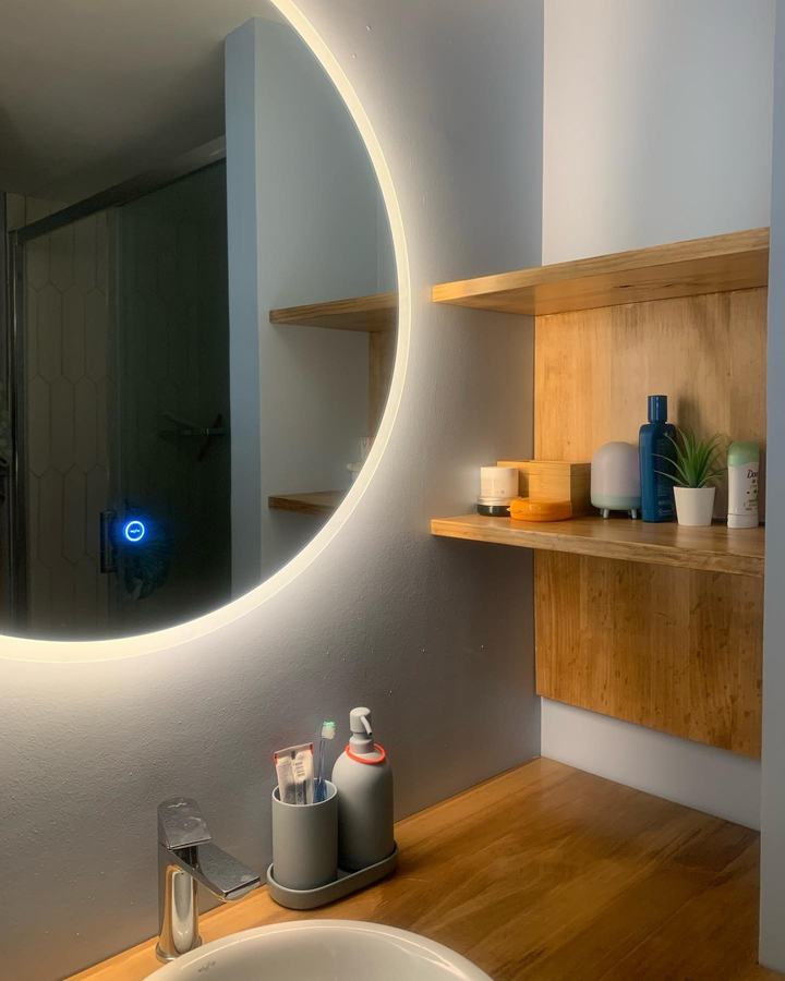 Baño con espejo circular retroiluminado y repisas de madera
